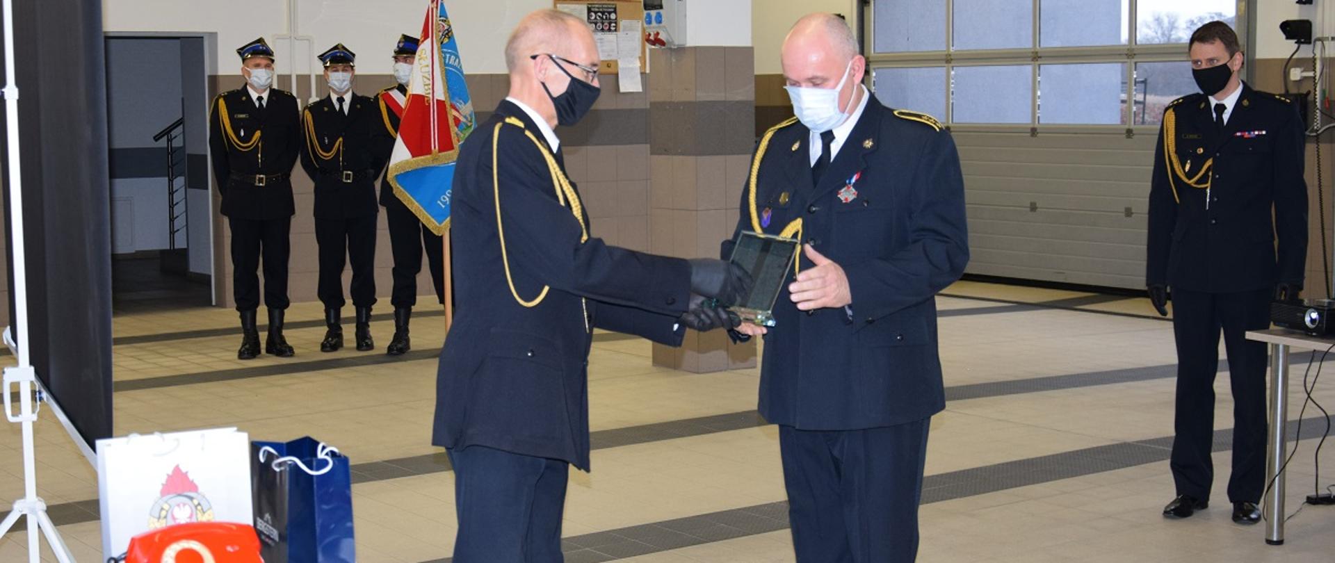 W pomieszczeniu garażowym JRG Wąbrzeźno Komendant Powiatowy Państwowej Straży Pożarnej st. bryg. Janusz Woźniak dziękuje asp. sztab. Pawłowi Makowskiemu Dyżurnemu Operacyjnemu Powiatu za służbę w Państwowej Straży Pożarnej. Obaj strażacy ubrani są w mundury wyjściowe. W tle stoi poczet sztandarowy wraz ze sztandarem komendy oraz dowódca uroczystości.