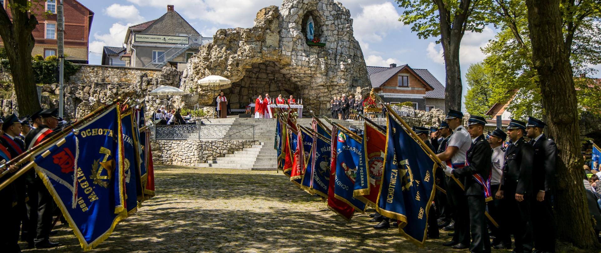 Poczty sztandarowe OSP i PSP w trakcie oddawaniu honoru w trakcie Mszy Świętej w grocie na Górze Świętej Anny. Uroczystość w ramach pielgrzymki strażaków. W tle kapłani w trakcie uroczystości.