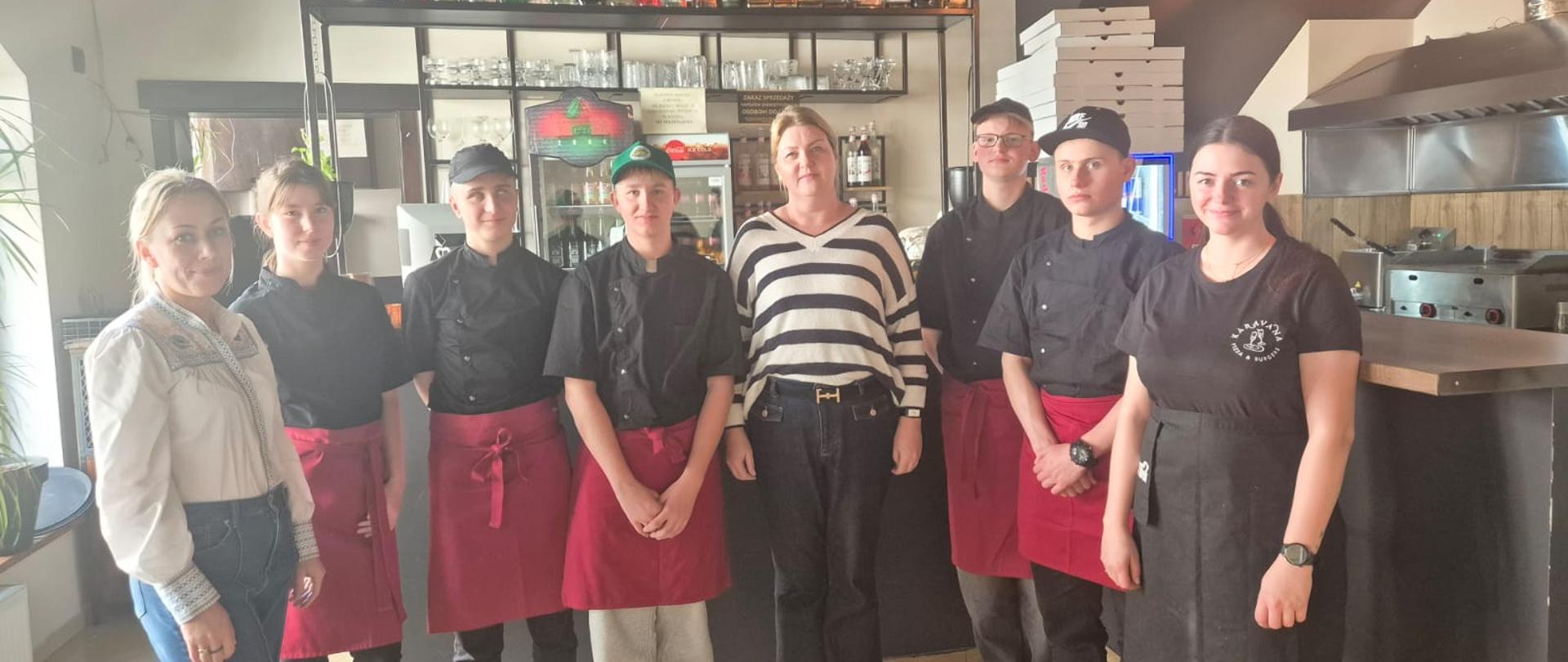 Zdjęcie przedstawia dwie Panie nauczycielki z przedmiotów zawodowych ZSCKR w Sypniewie wraz z uczniami o profilu technik żywienia i usług gastronomicznych podczas warsztatów KARAVANA PIZZA&BURGER w Tucholi pod opieką Pani Marii Żołnowskiej.