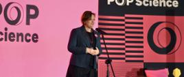 Zdjęcie z boku pod kątem, wiceminister Zioło-Pużuk w ciemnoszarej marynarce stoi na tle różowo-fioletowej ściany z napisem POP Science i mówi do mikrofonu na stojaku.
