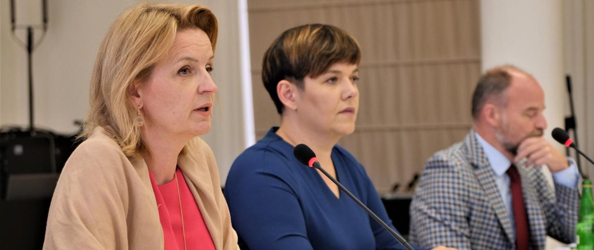 Zdjęcie przedstawia trzy osoby, siedzące obok siebie podczas spotkania pełnomocników. Jedna z nich przemawia do mikrofonu. 