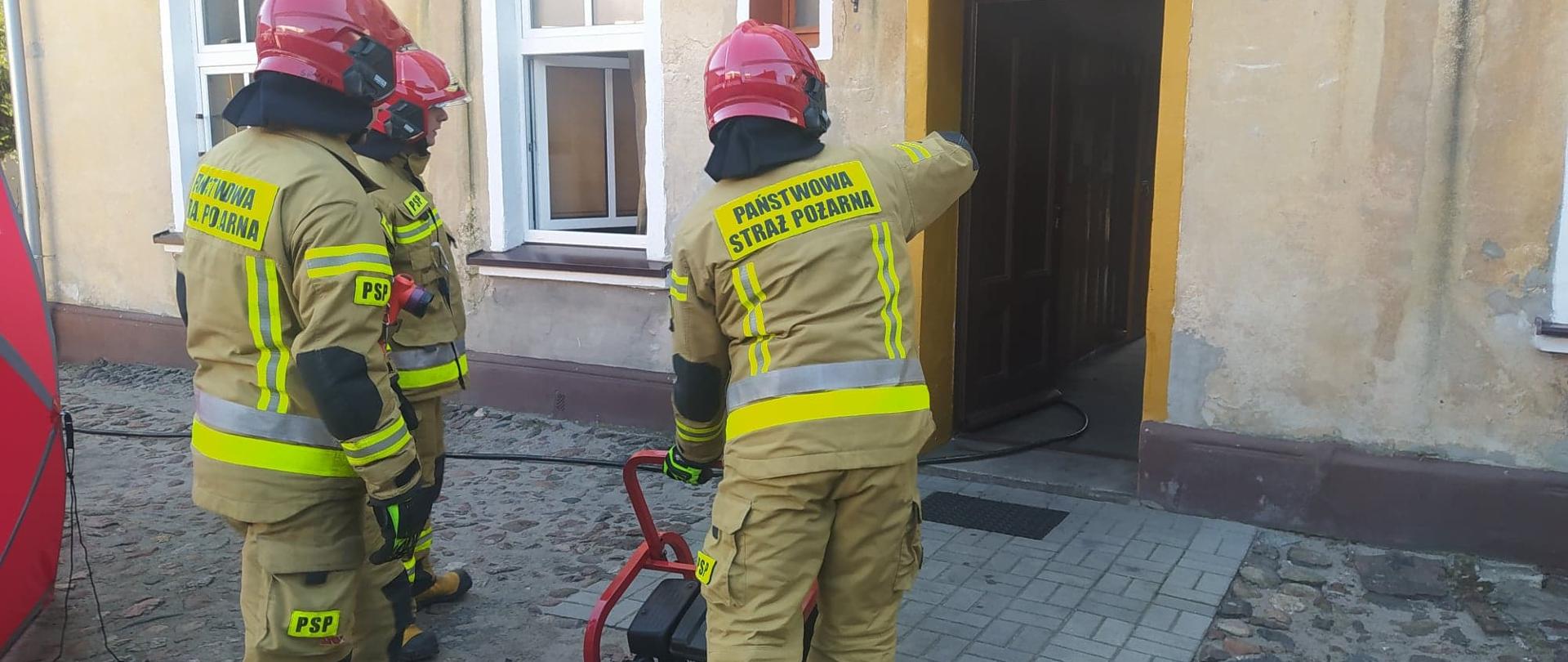 Tragiczny bilans pożaru. Posesja w Rawiczu przy ul. Piłsudskiego. Na podwórzu ustawiony jest parawan ochronny koloru czerwonego. Przed drzwiami wejściowymi do kamienicy znajdują się strażacy oraz wentylator nadciśnieniowy, przy pomocy którego prowadzone jest oddymianie i przewietrzanie budynku.