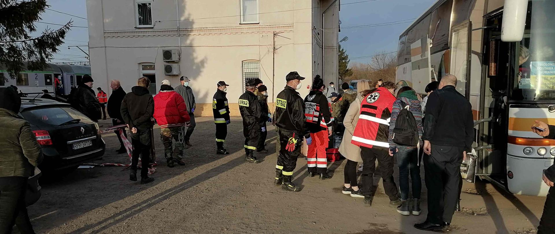 Na kamienistym utwardzonym placu przy autobusie stoi grupa osób: osoby cywilne, strażacy oraz ratownicy medyczni. Z tyłu duży budynek. Po prawej stronie czarny samochód osobowy, w tle fragment pociągu.