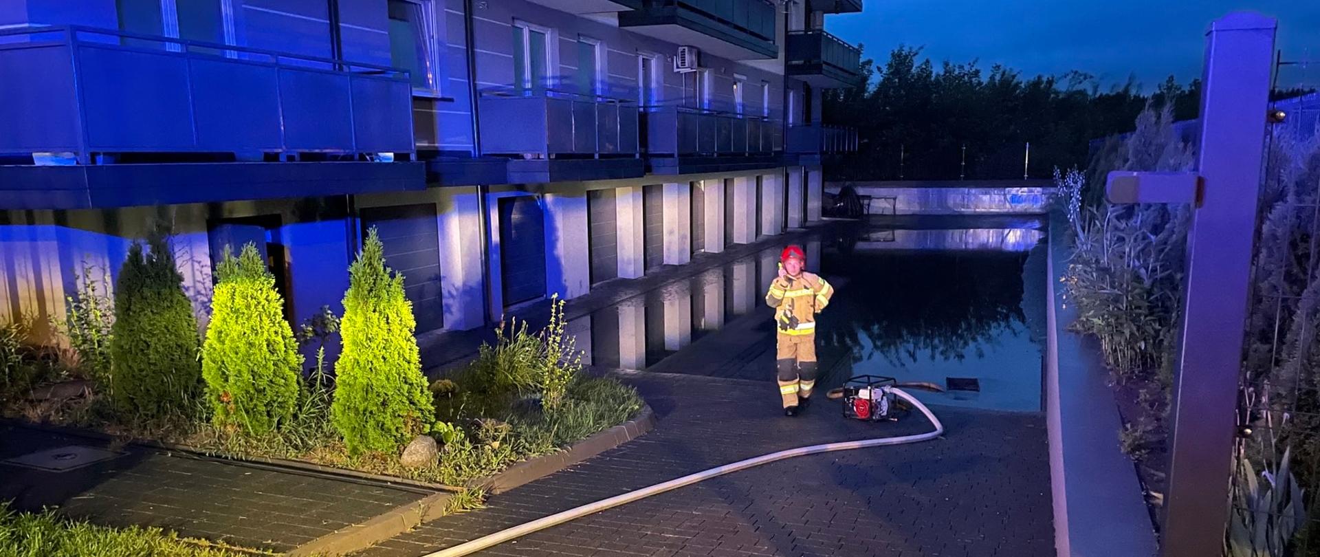 Noc, budynek mieszkalny-wielorodzinny, który posiada garaże w najniższej kondygnacji. Przed garażami widoczne rozlewisko, na skraju którego stoi pompa, a przy niej strażak. 