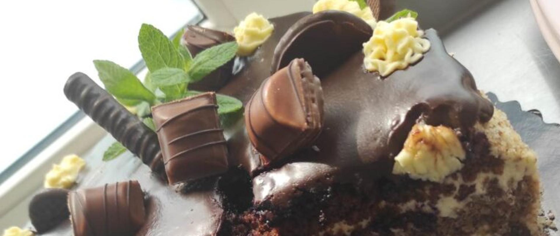 Okrągły tort z polewą czekoladową, z wyciętą ćwiartką, ciemne trzywarstwowe ciasta przełożone jasną masą. Ozdobiony słodyczami i jasną masą.