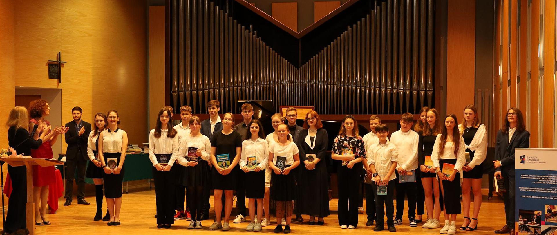 Zdjęcie na sali koncertowej. Na pierwszym planie publiczność. Uczniowie wyróżnienie stoją w dwóch rzędach trzymając w ręku nagrody książkowe. Z lewej strony stoi dyrekcja klaszcząc w dłonie. W tle za uczniami organy oraz fortepian, z lewej strony baner unijny.