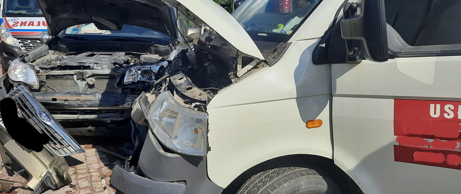 Na zdjęciu znajdują się dwa samochody po zderzeniu. Samochód dostawczy koloru białego ustawiony jest prostopadle do kierunku jazdy, pod nim posypana jest ulica sorbentem ponieważ wyciekł z niego olej silnikowy. Drugie auto osobowe koloru oliwkowego stoi wzdłuż kierunku jazdy, pod kątem prostym do auta dostawczego. Oba pojazdy mają otwarte maski i mają uszkodzone przody pojazdów. W tle za uszkodzonymi samochodami stoją karetki pogotowia ratunkowego.