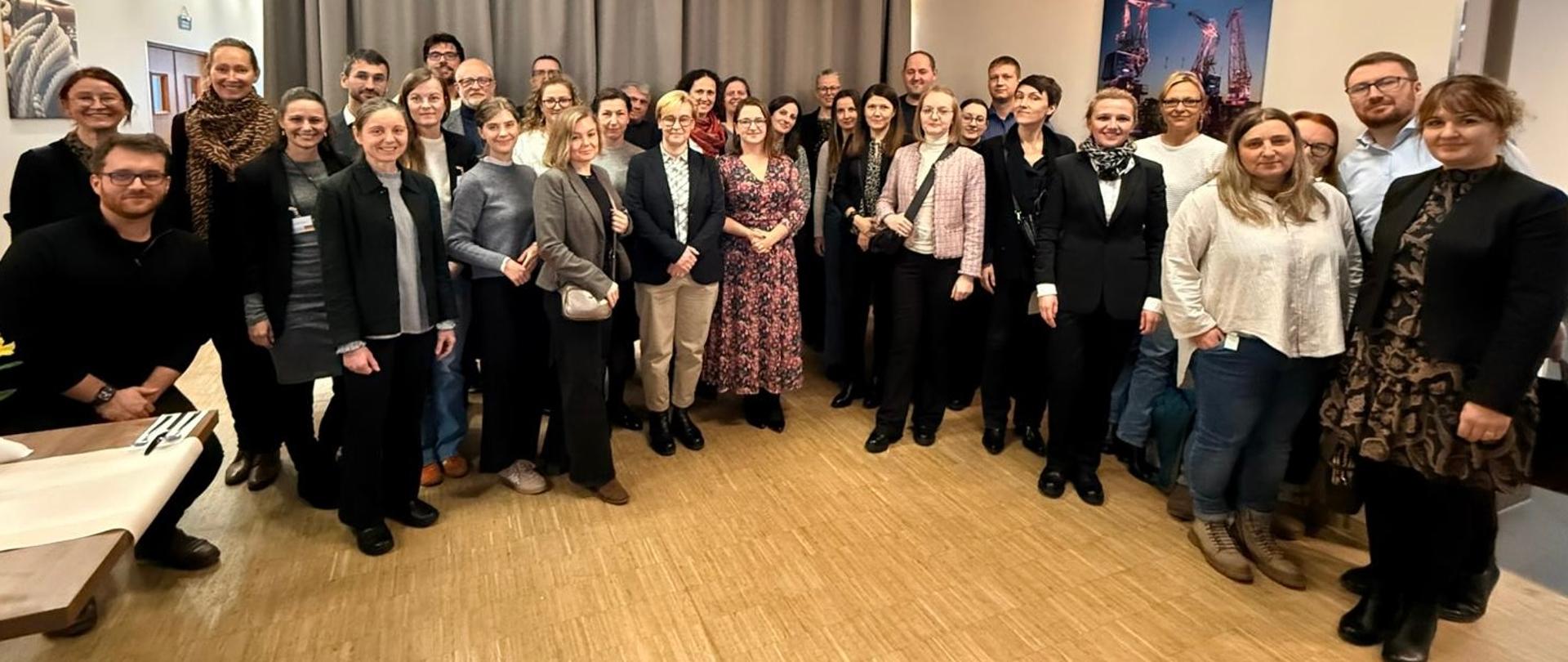 W sali w półokręgu stoi duża grupa osób, które się uśmiechają.