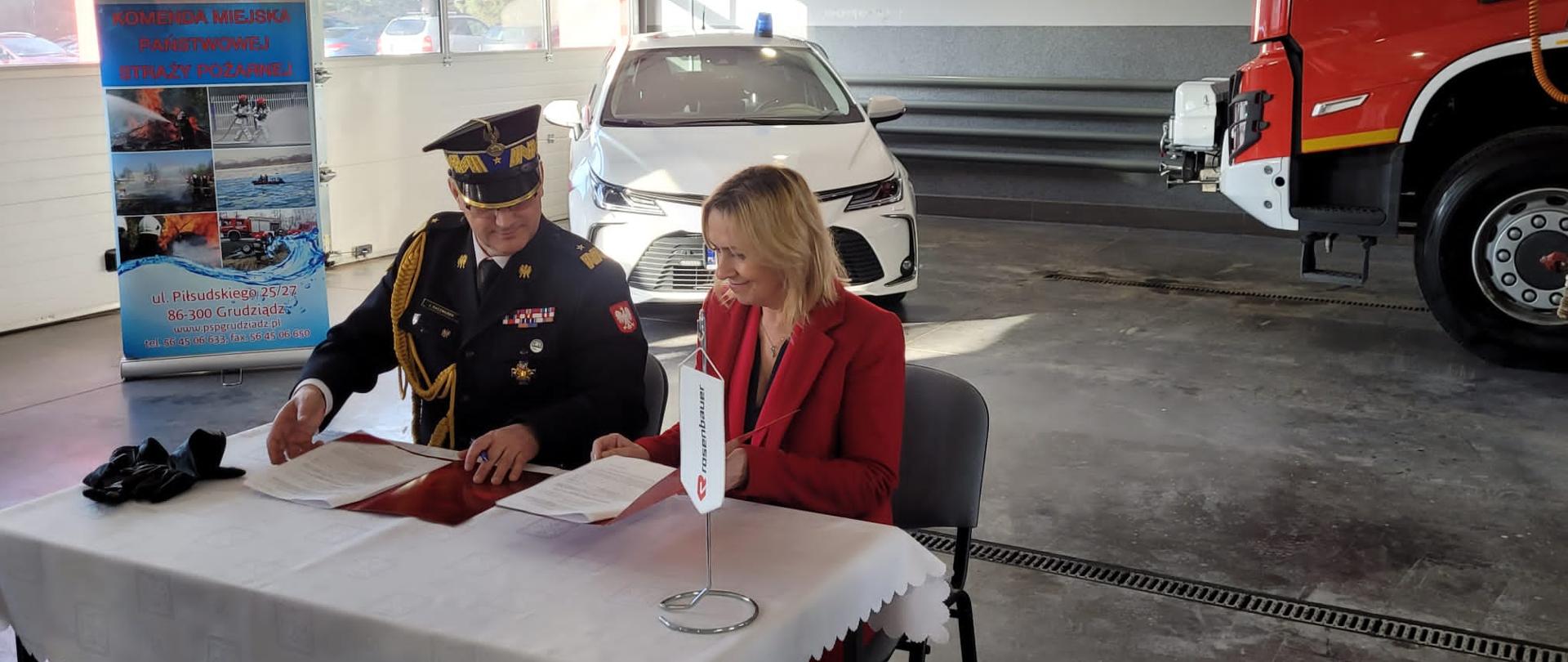 Przy stole siedzą komendant wojewódzki i przedstawiciel firmy Rosenbauer - kobieta. Podpisują umowę. W tle widać pojazdy. baner reklamowy, wrota garażowe.