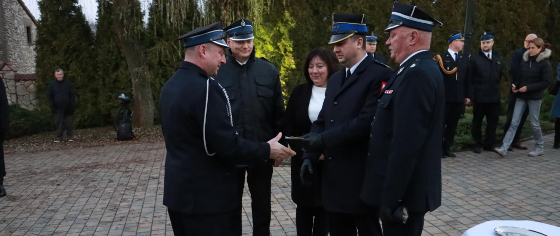Zdjęcie przedstawia moment wręczenia kluczyków do nowego samochodu ratowniczo-gaśniczego. Przekazania dokonuje funkcjonariusz PSP w asyście druhów OSP i kobiety w ubiorze cywilnym. W tle znajdują się inni druhowie i funkcjonariusze.