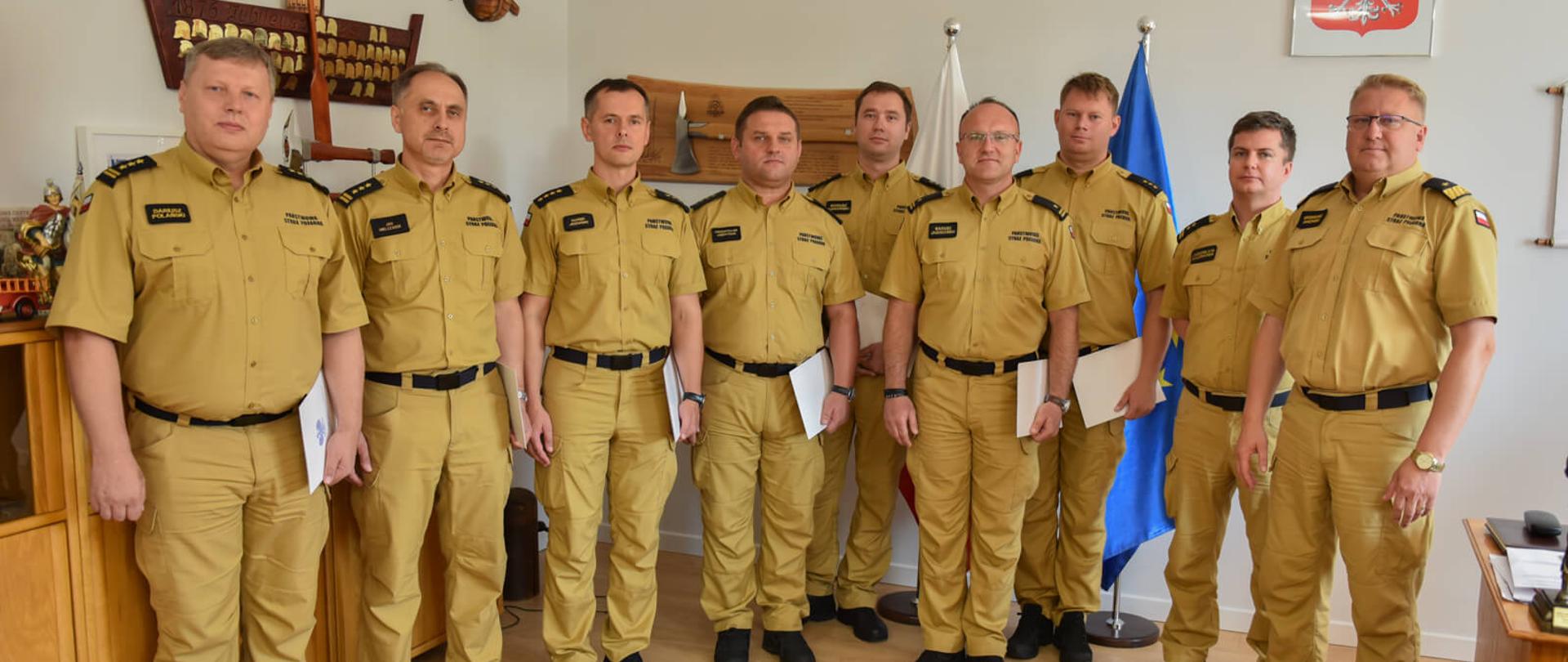 dziewięciu strażaków w piaskowych mundurach, w koszulach z krótkim rękawem pozuje do zdjęcia w sali, na sianach różne elementy ozdobne z motywami strażackimi 