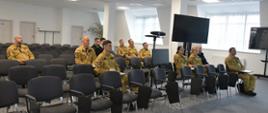 Strażacy w mundurach koloru piaskowego siedzą na krzesłach obok nich stoją monitory.