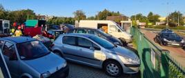 Kilka samochodów osobowych w tym jeden z przyczepką stojących na ogrodzonym parkingu przyległym do drogi. Za samochodami po lewej stronie widoczna grupa stojących strażaków i gapiów. Strażacy trzymają parawan. Przed parawanem dwóch klęczących strażaków oraz pochylony ratownik medyczny. Poszkodowany niewidoczny na zdjęciu, zasłonięty przyczepką przypiętą do szarego samochodu osobowego.