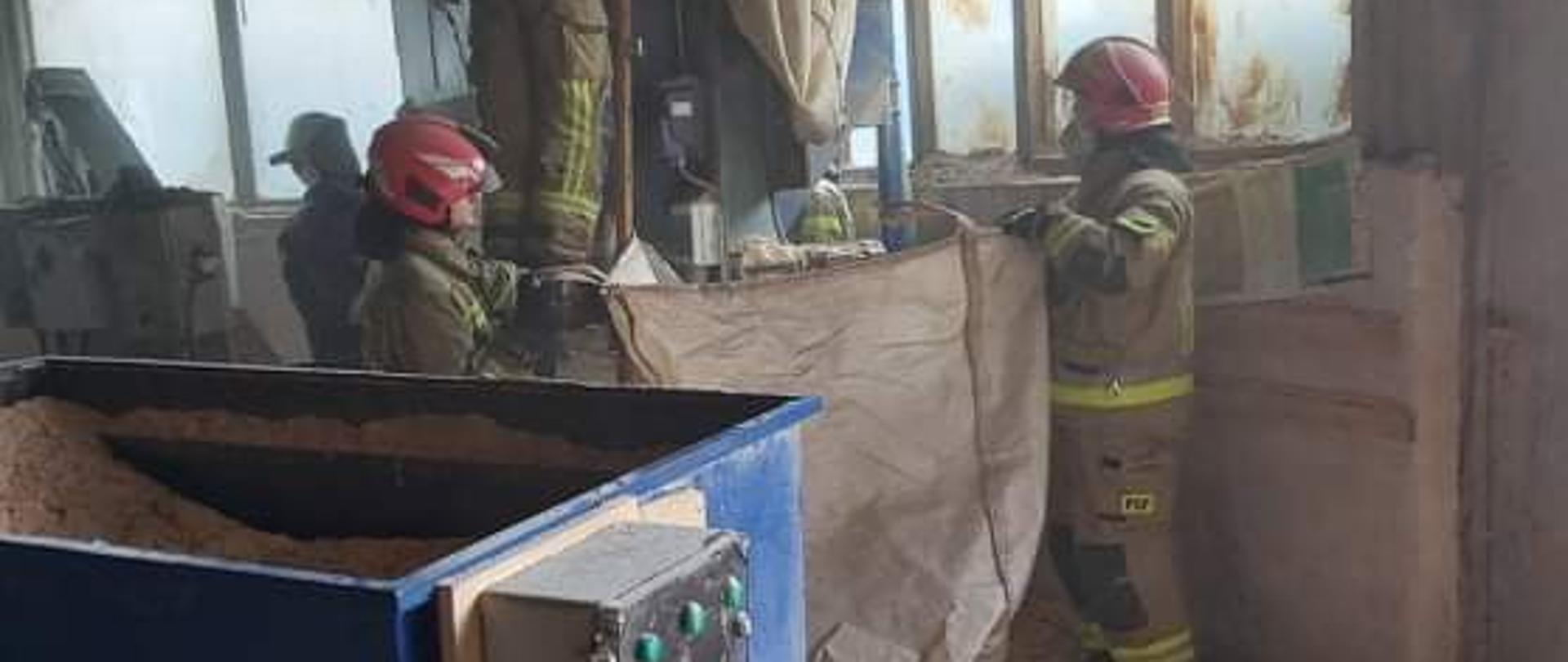 Na pierwszym planie zdjęcia widoczny jest niebieski zbiornik, do którego na jednej ze ścianek zamontowane są dwa silniki, jeden w kolorze szarym, a drugi w kolorze zielonym. Na tej samej ściance znajduje się również panel z przyciskami sterującymi w kolorach czarnym, zielonym i czerwonym. W zbiorniki znajdują się trociny. Na podłodze budynku znajdują się rozsypane trociny. W dalszej części zdjęcia widać czterech strażaków w specjalnych ubraniach, którzy gaszą palące się trociny w drugim ze zbiorników. Dwóch strażaków trzyma duży biały worek , a trzeci znajduje się na podeście zbiornika i zalewa wodą palące się trociny. W tle za zbiornikiem widać stojącego czwartego strażaka biorącego udział w zdarzeniu. Pomieszczenie jest lekko zadymione. Na dwóch ścianach budynku znajdują się duże okna. 