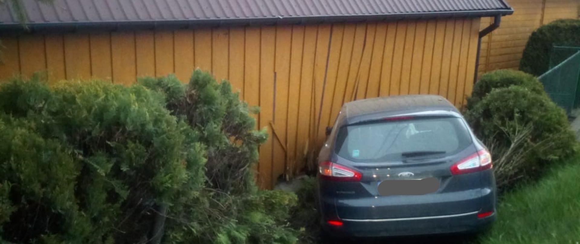 Na zdjęciu widoczny jest samochód osobowy, który uderzył w budynek gospodarczy- uszkodzenia drewnianej ściany. Poniżej widzimy zniszczona siatkę ogrodzeniową oraz krzewy.
