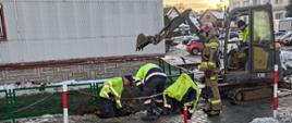 Widok z boku. Likwidacja zagrożenia związanego z wyciekiem gazu. Po prawej stronie minikoparka. Pracownicy pogotowia gazowego nad wykopem z łopatami. 