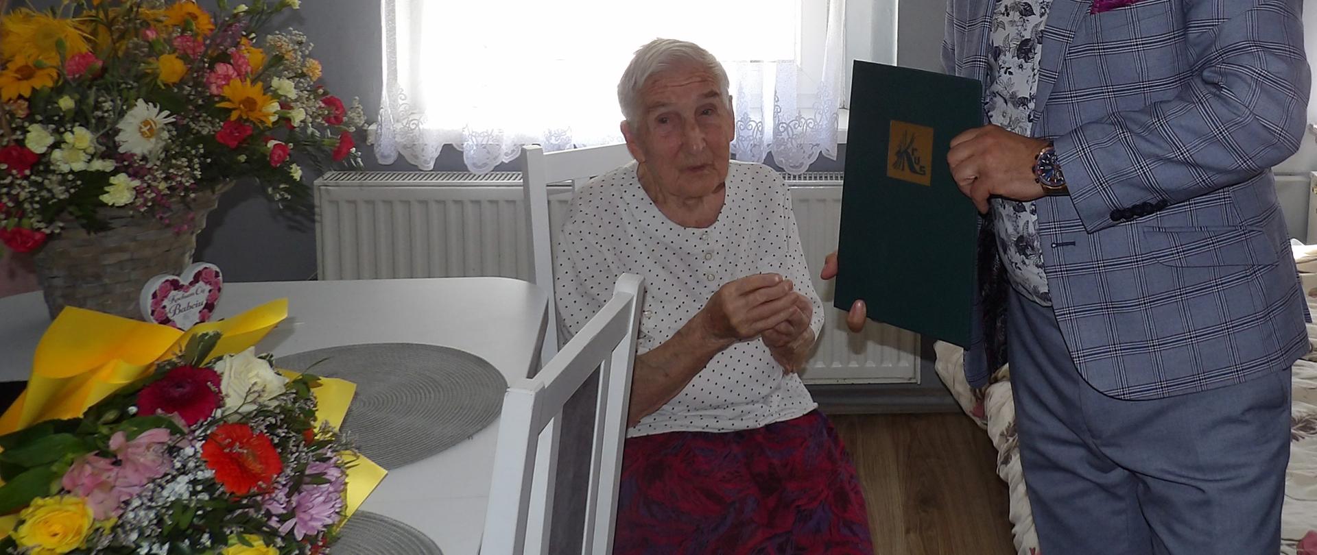 W niedużym pokoju przy oknie na krześle siedzi starsza kobieta - Jubilatka. Obok niej, po prawej stronie, stoi mężczyzna - kierownik PT KRUS w Lubaniu, ubrany w szarą marynarkę w kratkę w rękach trzyma ozdobny zielony dyplom ze złotym logo KRUS.