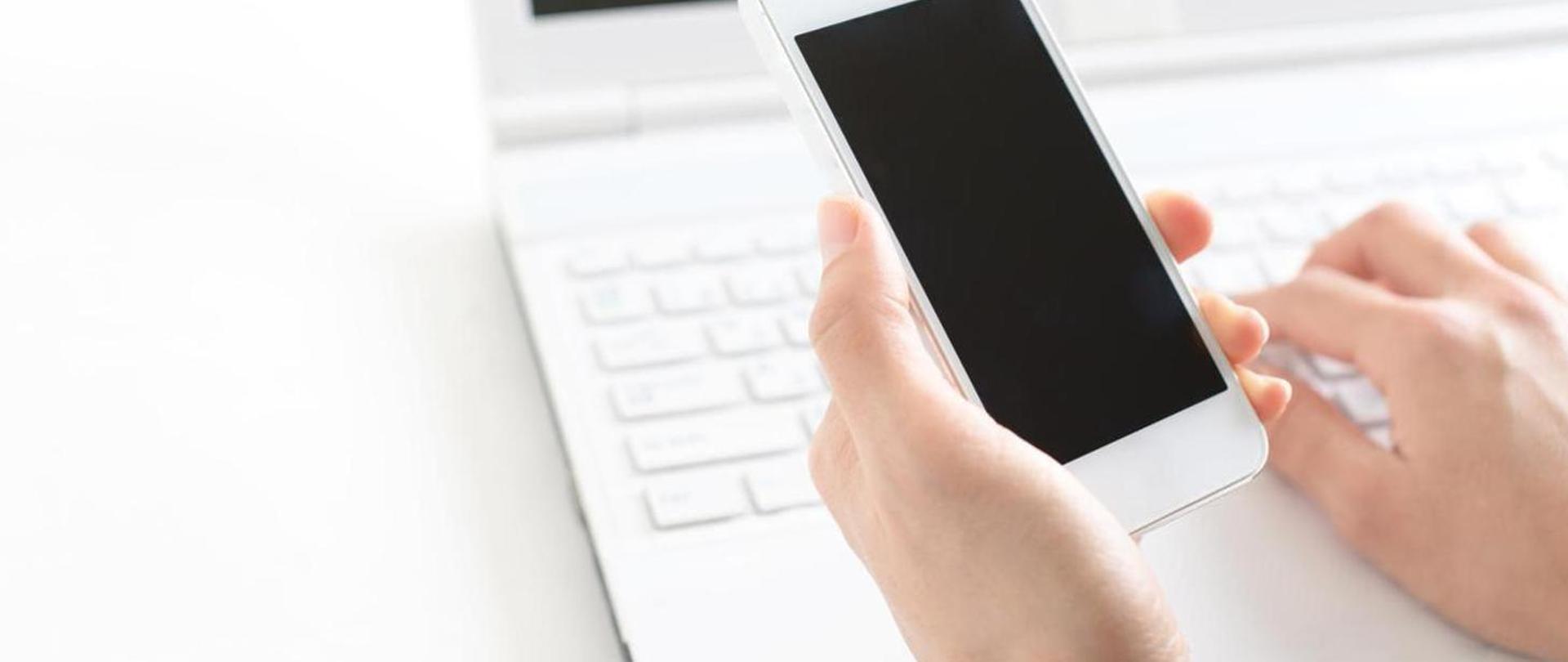na zdjęciu dwie dłonie z smartfonem, w tel zdjęcia biały laptop. 