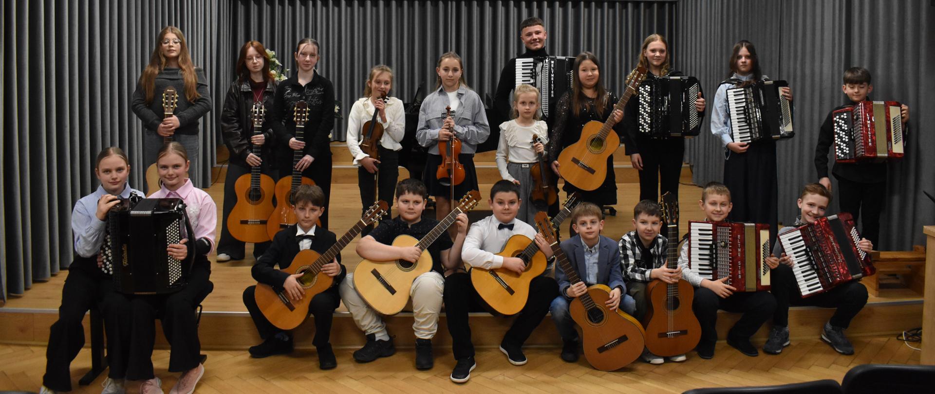 Zdjęcie przedstawia aulę państwowej szkoły Muzycznej I st. im. Witolda Lutosławskiego w Jaśle. Ściany auli posiadają kolor grafitowy, a podłoga jasnobrązowy. Na tylnej ścianie znajduje się logo szkoły oraz godło Polski. Na głównym planie widać uczniów klasy gitary Jarosława Stompla, skrzypiec Anny Cieślickiej oraz akordeonu Jakuba Zastawnego. Uczniowie pozują do zdjęcia na scenie auli. Część z nich siedzi na krzesłach lub podeście. Reszta uczniów stoi w trzecim rzędzie. Wszyscy wykonawcy trzymają w rękach instrument na którym uczą się gry w szkole.