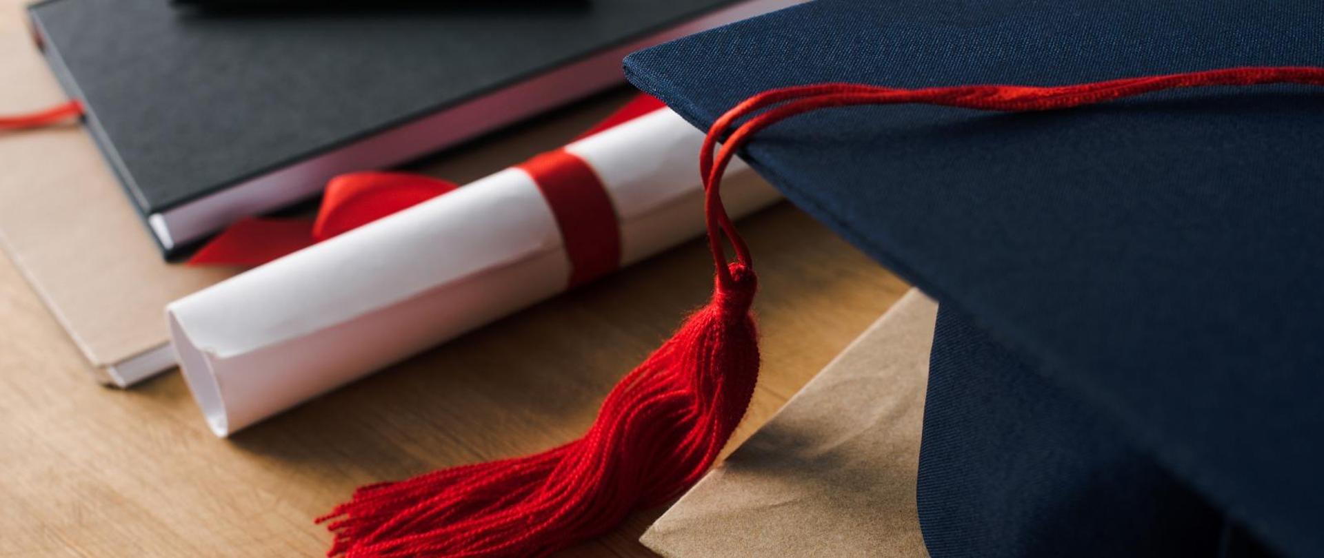 Grafika - na stole leży studencki biret, dwie książki oraz związany tasiemką dokument.