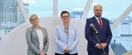 Pod półokrągłym oknem stoi wiceminister Mrówczyńska, wiceminister Lubnauer i dyrektor IBE w granatowym garniturze.