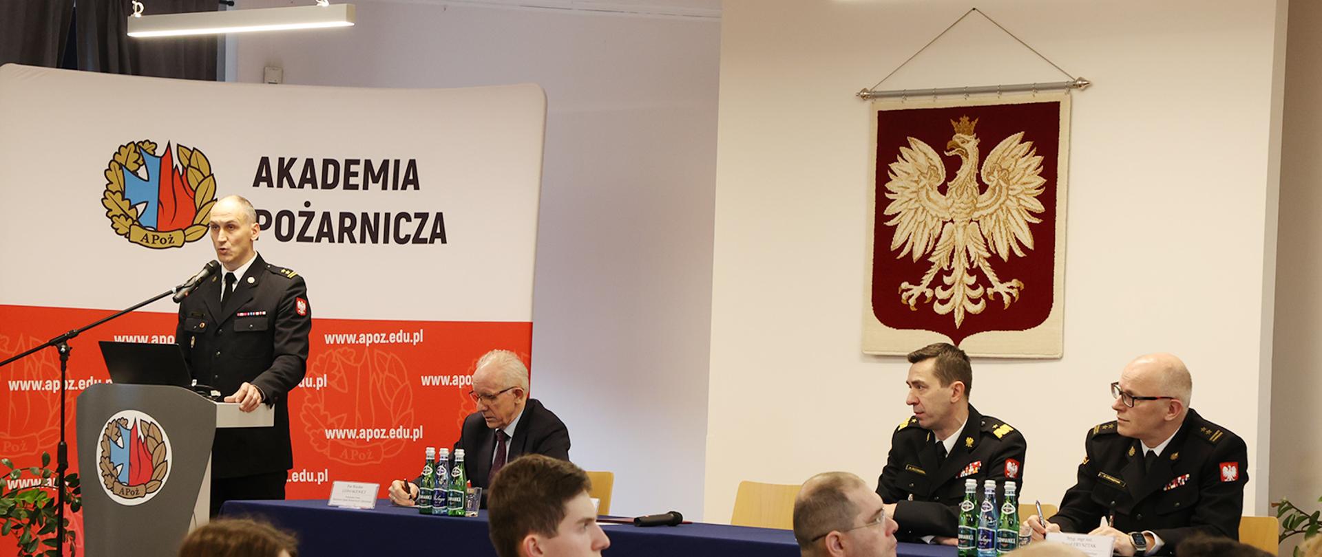 Do mikrofonu przemawia rektor Akademii, obok przy stole siedzi wiceminister SWiA oraz komendant główny PSP i zastępca, przed nimi siedzą uczestnicy narady