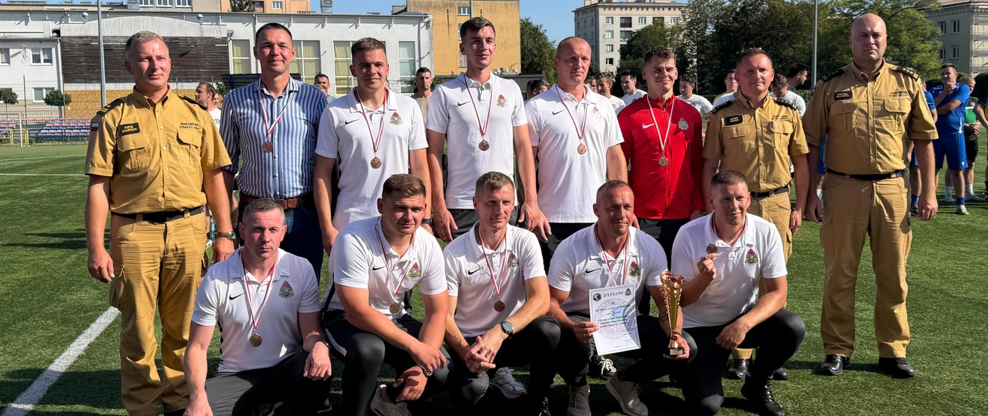 Opis: Zbiorowe zdjęcie reprezentacji / zespołu wraz z zaproszonymi gośćmi oraz przedstawicielami Państwowej Straży Pożarnej po ceremonii dekoracji.
Szczegóły:
Zawodnicy mają na sobie białe koszulki polo (oraz czerwoną bluzę) i wyeksponowane na szyi medale.
Jeden z zawodników na dole trzyma oficjalny dyplom i puchar.
Zespół otoczony jest przez funkcjonariuszy PSP w mundurach służbowych.
Wszystkim dopisują wyśmienite humory, a uroczystość odbywa się przy pełnym słońcu na boisku w Radomiu.