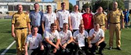 Opis: Zbiorowe zdjęcie reprezentacji / zespołu wraz z zaproszonymi gośćmi oraz przedstawicielami Państwowej Straży Pożarnej po ceremonii dekoracji.
Szczegóły:
Zawodnicy mają na sobie białe koszulki polo (oraz czerwoną bluzę) i wyeksponowane na szyi medale.
Jeden z zawodników na dole trzyma oficjalny dyplom i puchar.
Zespół otoczony jest przez funkcjonariuszy PSP w mundurach służbowych.
Wszystkim dopisują wyśmienite humory, a uroczystość odbywa się przy pełnym słońcu na boisku w Radomiu.