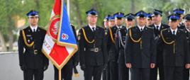 Grupa strażaków w granatowych mundurach. Od lewej poczet sztandarowy.