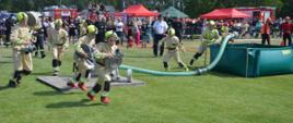 Zawody sportowo-pożarnicze. Płyta boiska na stadionie sportowym. Ćwiczenie bojowe wykonuje jedna z MDP. Zawodnicy z weżami i innym sprzetem w rękach biegną w stronę "pożaru". W tle publiczność, namioty i wozy strażackie.