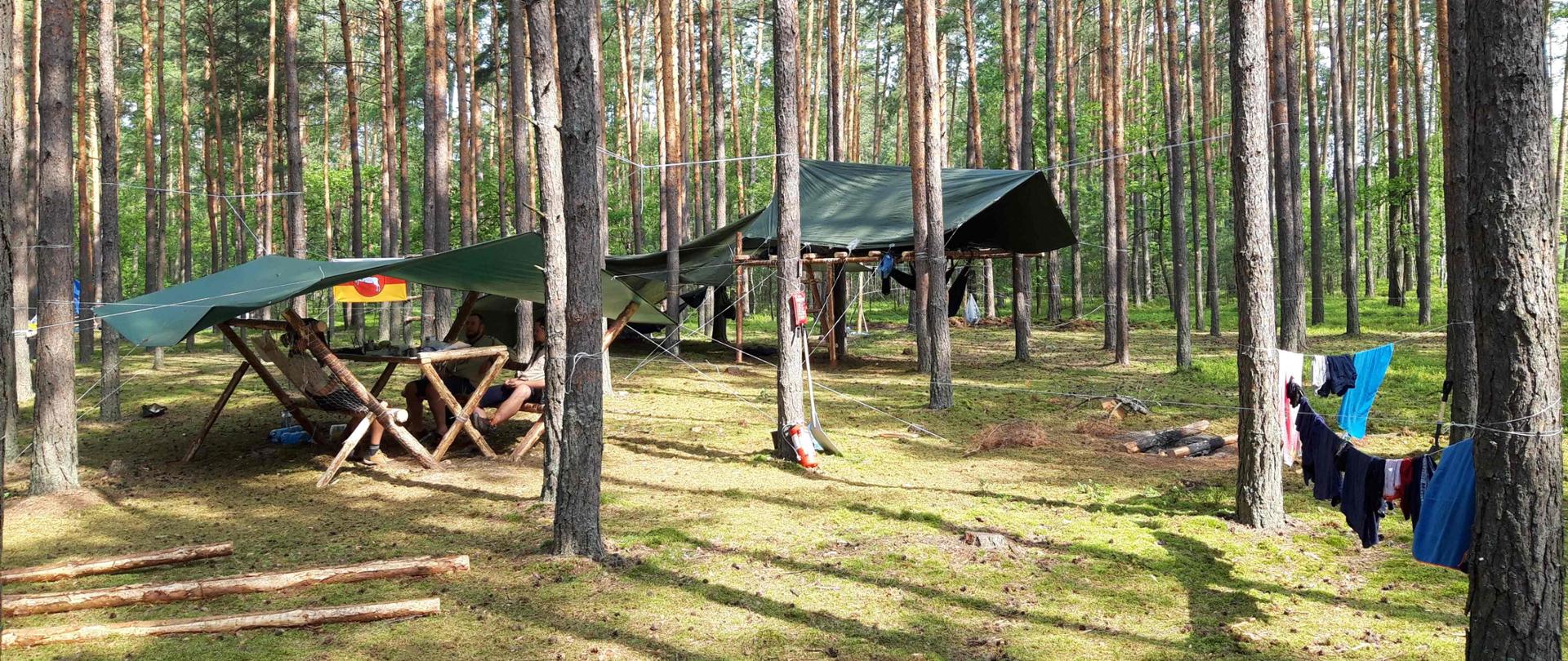 Na zdjęciu obóz harcerski w lesie. Po lewej stronie zadaszenie z plandeki zawieszonej pomiędzy drzewami przy użyciu sznurków, pod nim stół oraz siedziska z oparciem wykonane ze stempli, przy których siedzą 3 osoby. Po prawej w oddali kolejne zadaszenie z plandeki, pod nim konstrukcja ze stempli. Zdjęcie wykonane w słoneczny dzień.