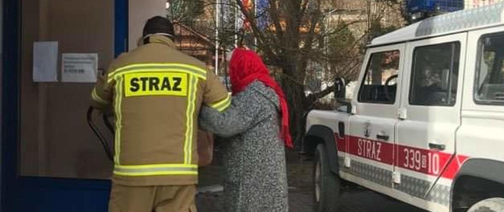 Zdjęcie prezentuje strażaka w żółtym ubraniu specjalnym zrobione od tyłu , który pod rękę pomaga starszej kobiecie w czerwonej chuście na głowie i szarym płaszczu dotrzeć do punktu szczepienia. Z prawej strony zdjęcia samochód strażacki koloru białego z napisem STRAŻ na czerwonym pasie