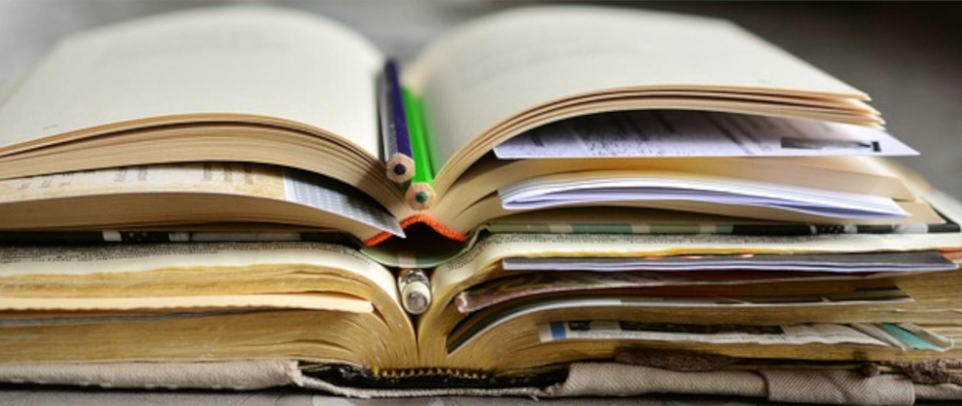 Fotografia przedstawiająca stertę otwartych książek poprzekładaną ołówkami i kartkami z notatkami.