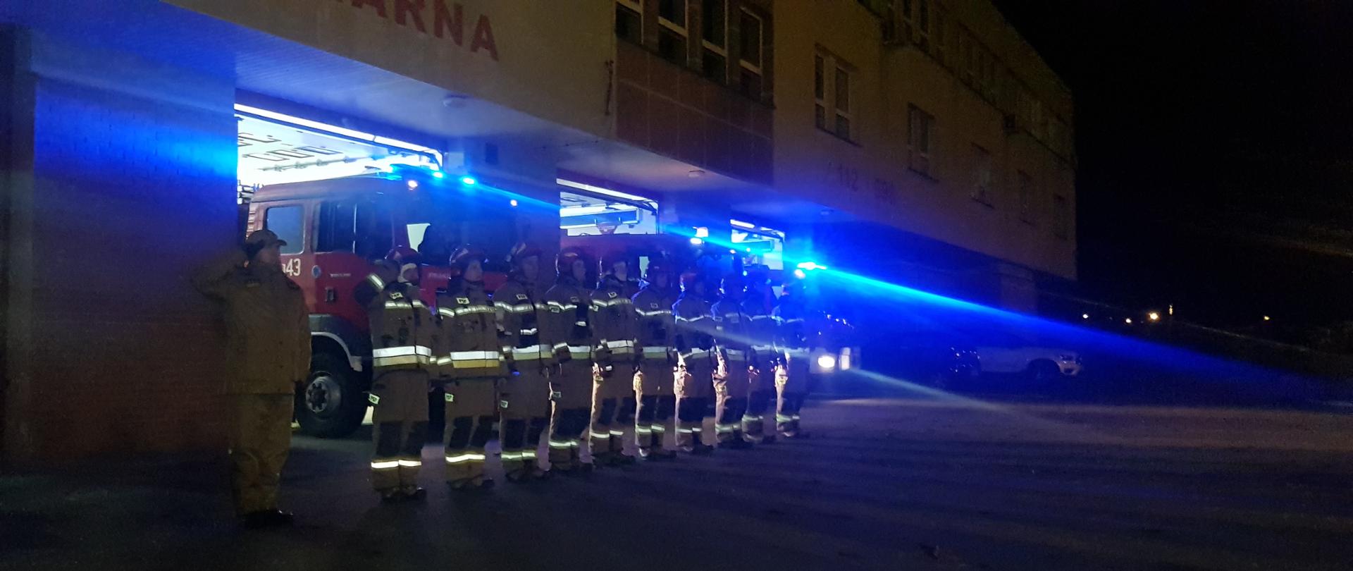 Strażacy przed strażnicą oddają hołd dla druhów z Czernikowa przed garażami. Strażacy w pełnym umundurowaniu bojowym. W tle trzy samochody ratowniczo - gaśnicze na sygnałach świetlnych