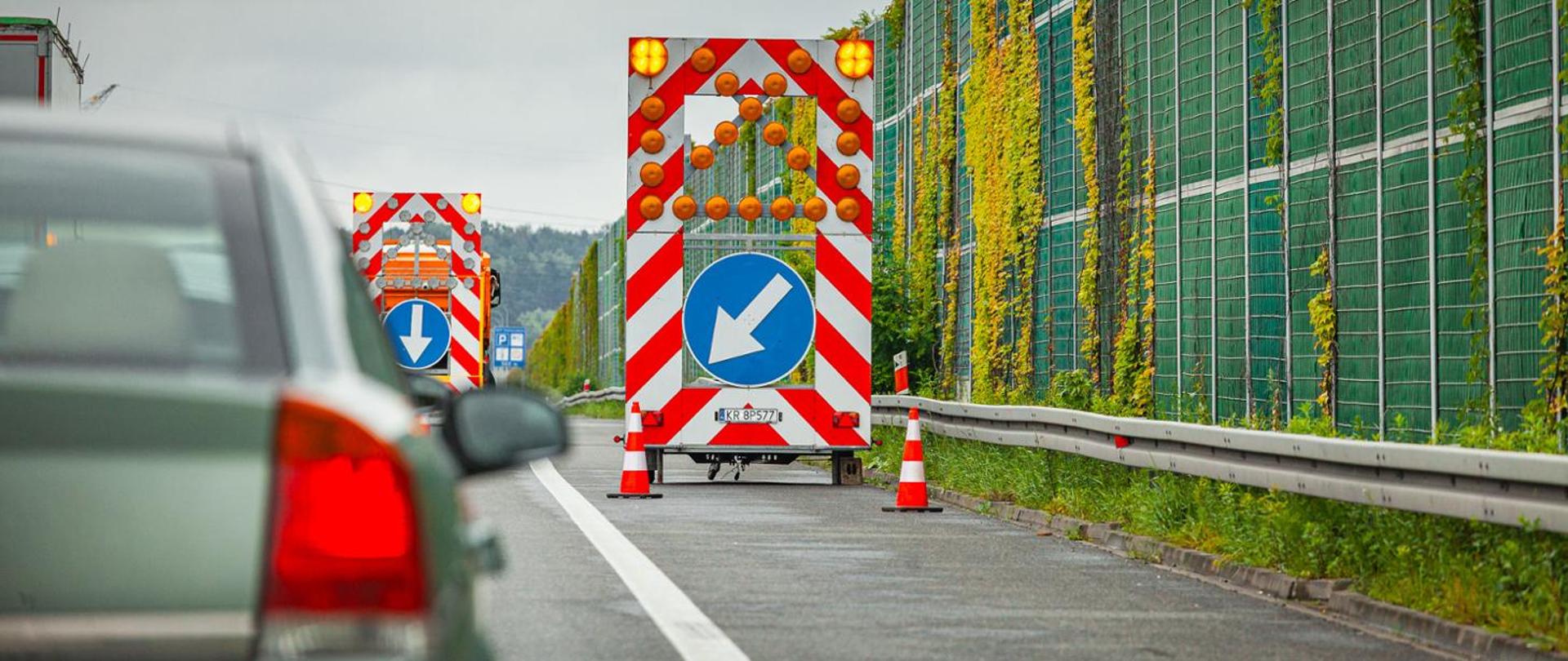 Prace drogowe na autostradzie A1. Na pasie awaryjnym ustawiono pojazdy z tablicami świetlnymi ostrzegającymi kierowców o zwężeniu jezdni i konieczności zmiany toru jazdy. Obok przebiegają ekrany akustyczne porośnięte pnączami, a w kadrze widoczny jest również przejeżdżający samochód.