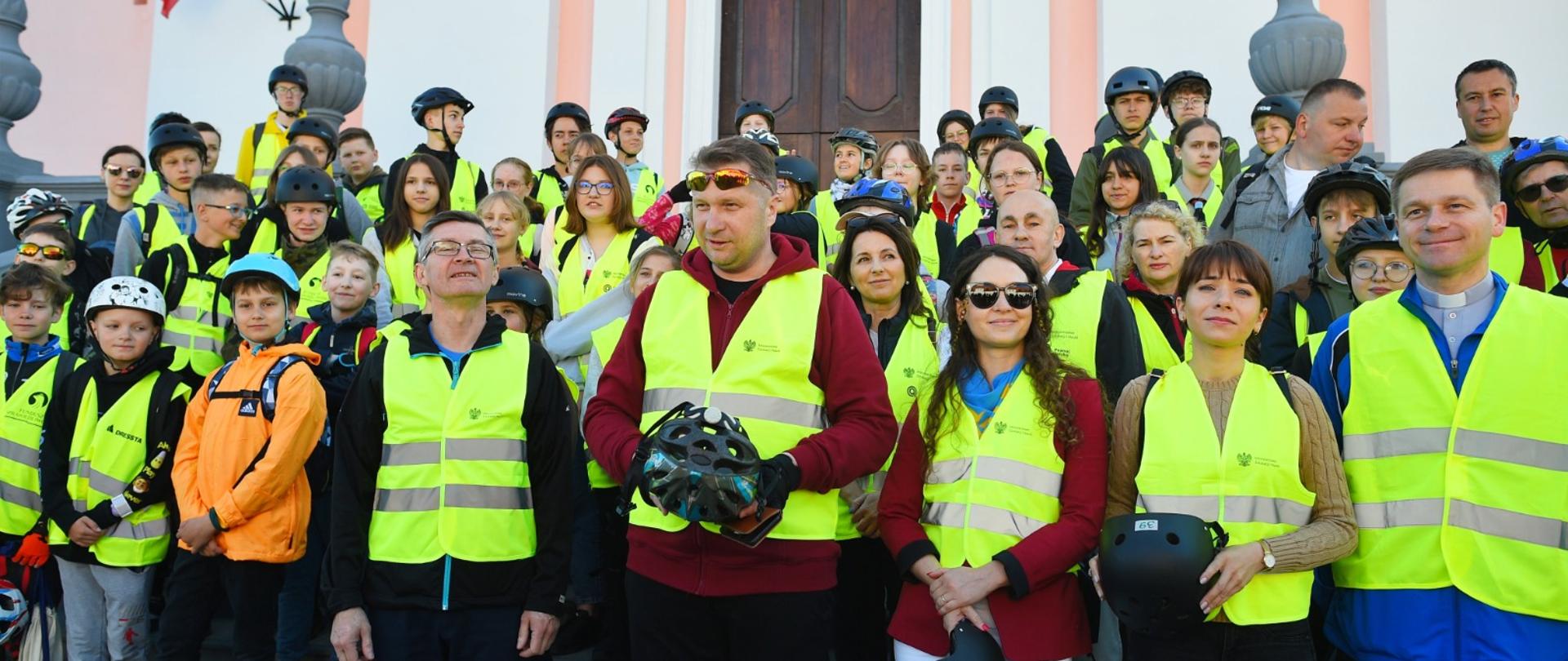 Minister Czarnek w żółtej kamizelce stoi pośrodku dużej grupy osób w takich samych kamizelkach, w tle budynek z biało-różowymi ścianami.