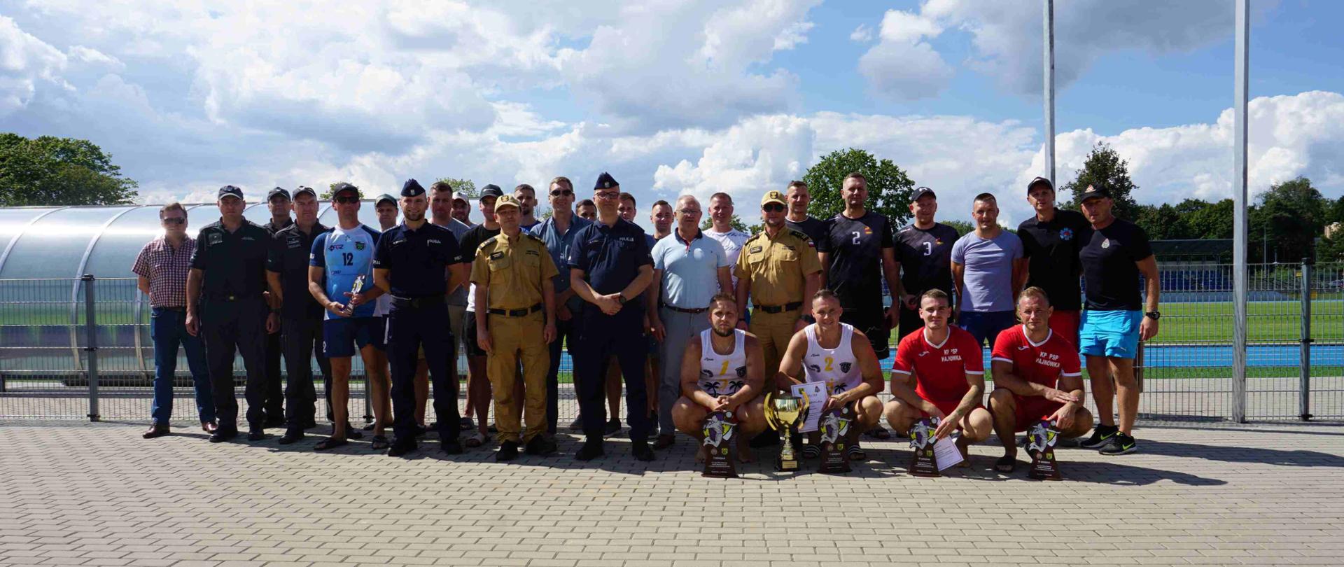 Na chodniku w dwuszeregu stoi kilkadziesiąt mężczyzn. Przed kilkoma, którzy kucnęli stoją puchary. 