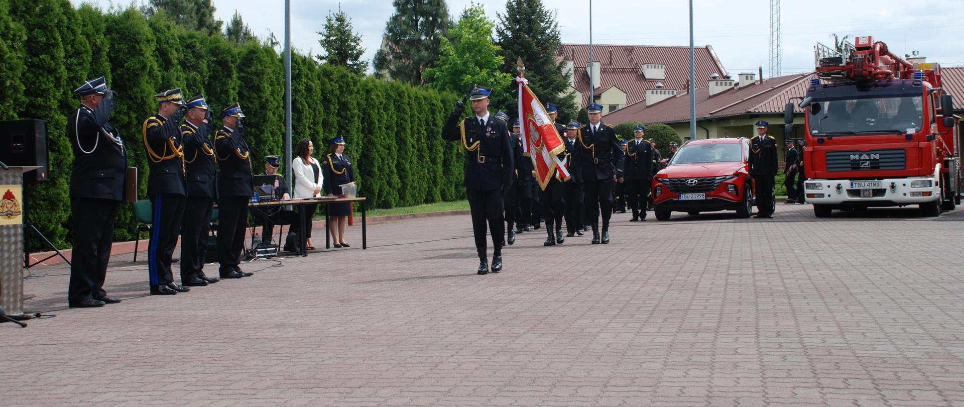 Po lewej stronie stoi 4 strażaków w mundurach galowych i czapkach, przy których daszku mają 2 palca. Na prosto idzie defilada, na przodzie dowódca uroczystości w mundurze, a za nim po 3 osoby obok siebie za sztandarami idą pozostali strażacy. Na prawo w tle widać 2 samochody pożarnicze. W tle również budynki.