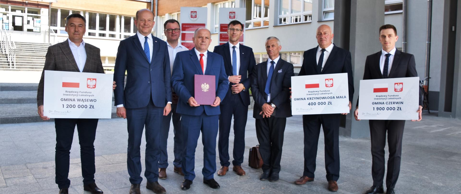 Na zdjęciu znajduje się 8 elegancko ubranych mężczyzn. Większość z nich trzyma w rękach symboliczne promesy ze środkami wsparcia finansowego z Rządowego Funduszu Inwestycji Lokalnych. 