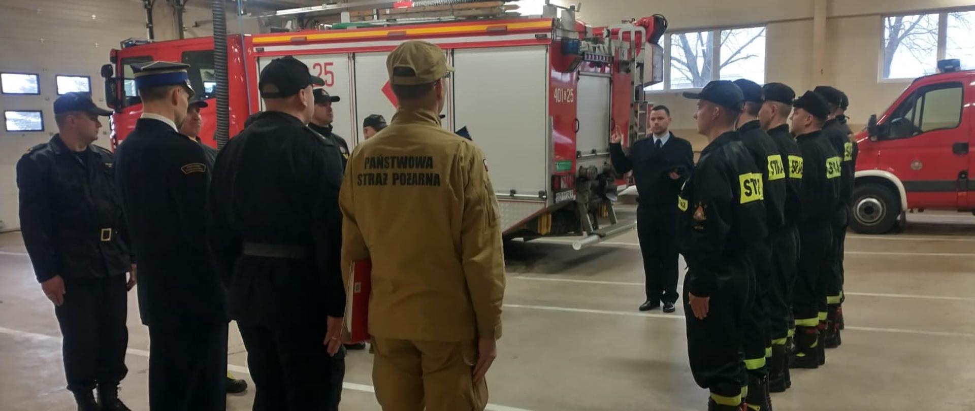 Zdjęcie zostało zrobione w garażu PSP. Przedstawia ślubowanie nowego funkcjonariusza. Funkcjonariusz w mundurze galowym stoi naprzeciwko Komendanta PSP oraz Dowódców. Ma uniesioną prawą rękę, a w lewej trzyma czapkę. Po jego prawej i lewej stronie stoją w rzędzie strażacy zmiany. W tle widać dwa wozy strażackie.