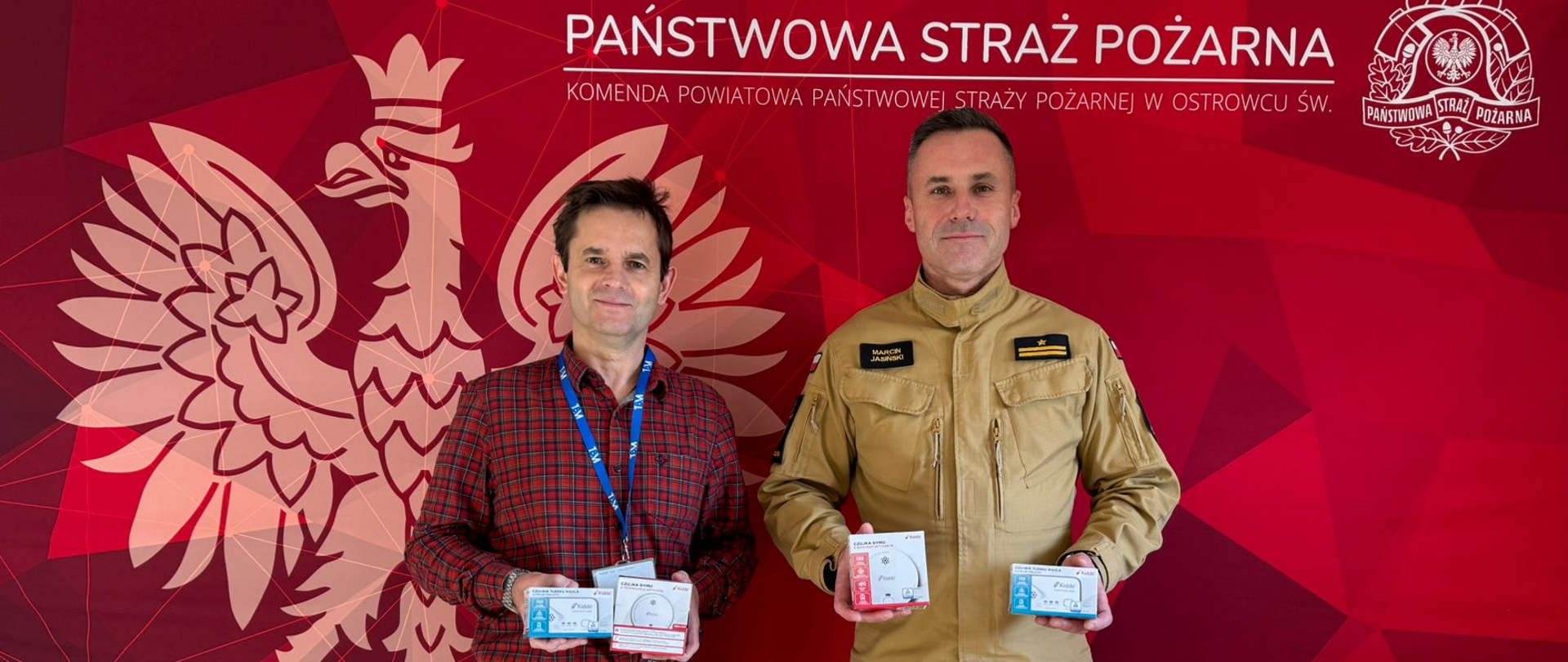 Na zdjęciu widoczny jest Komendant Powiatowy Państwowej Straży Pożarnej w Ostrowcu Św. oraz przedstawiciel przedsiębiorstwa TBM SNACKS Sp. z o. o. w rękach trzymają czujki dymu i tlenku węgla.