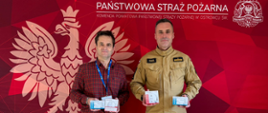 Na zdjęciu widoczny jest Komendant Powiatowy Państwowej Straży Pożarnej w Ostrowcu Św. oraz przedstawiciel przedsiębiorstwa TBM SNACKS Sp. z o. o. w rękach trzymają czujki dymu i tlenku węgla.
