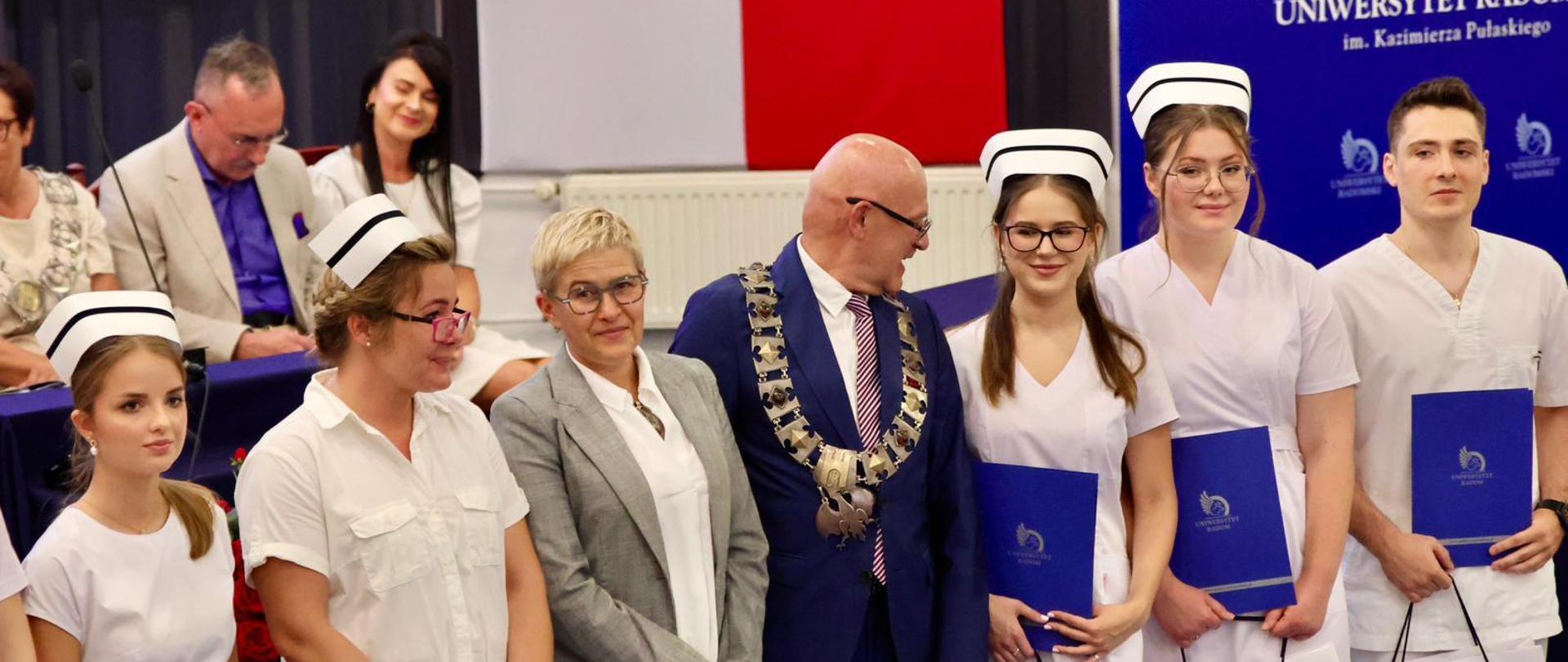 Na sali stoi szereg młodych kobiet w białych fartuchach i pielęgniarskich czepkach, między nimi wiceminister Mrówczyńska i mężczyzna w granatowym garniturze.