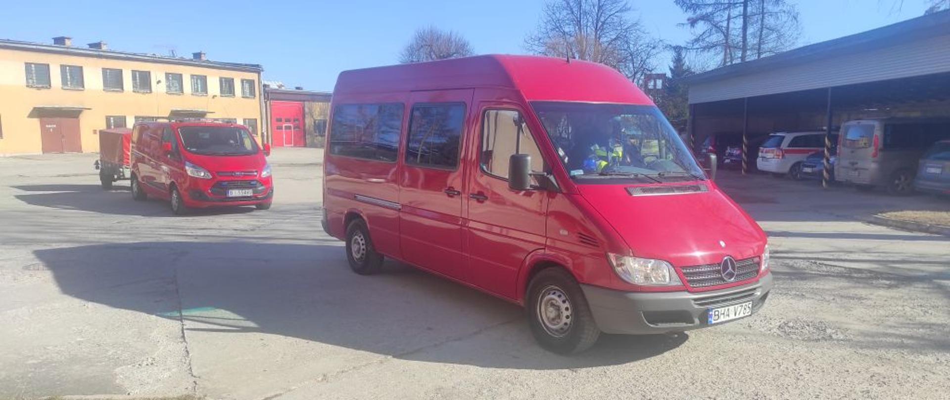 Na placu o betonowej nawierzchni przejeżdżają dwa czerwone strażackie busy. Do jednego zaczepiona jest czerwona przyczepka.