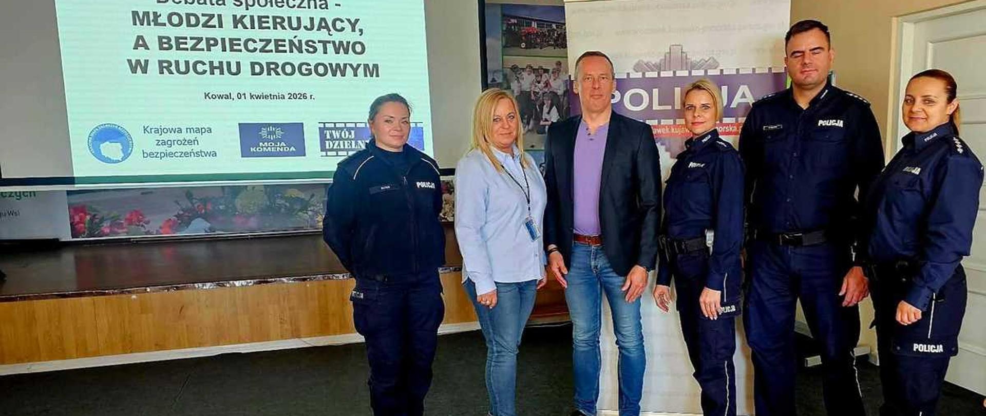 Zdjęcie przedstawia grupę sześciu osób pozujących do wspólnej fotografii po zakończeniu debaty społecznej. Spotkanie odbywa się w sali konferencyjnej, prawdopodobnie w Kowalu. W grupie znajduje się czworo funkcjonariuszy policji w pełnym umundurowaniu oraz dwoje cywilów.
Postacie i układ (od lewej do prawej)
Policjantka: Kobieta o jasnych, spiętych włosach, ubrana w granatowy mundur policyjny.
Kobieta (cywil): Ma długie, blond włosy, ubrana jest w jasnoniebieską koszulę i jeansy.
Mężczyzna (cywil): Wysoki mężczyzna w ciemnej marynarce, fioletowym t-shircie i jeansach. Stoi w centrum grupy.
Druga policjantka: Kobieta o jasnych włosach, w granatowym mundurze.
Policjant: Wysoki, postawny mężczyzna w granatowym mundurze policyjnym.
Trzecia policjantka: Kobieta o ciemnych włosach spiętych w kucyk, również w pełnym umundurowaniu.
Tło i ważne detale
Ekran projekcyjny: Po lewej stronie, w głębi, widać duży ekran z wyświetloną prezentacją. Napis na slajdzie głosi: „Debata społeczna – MŁODZI KIERUJĄCY, A BEZPIECZEŃSTWO W RUCHU DROGOWYM. Kowal, 01 kwietnia 2026 r.”. Na dole slajdu widoczne są logotypy akcji: „Krajowa mapa zagrożeń bezpieczeństwa”, „Moja Komenda” oraz „Twój Dzielnicowy”.
Banner: Po prawej stronie za grupą stoi biało-fioletowy roll-up z dużym napisem „POLICJA” oraz logo Komendy Powiatowej Policji.
Sala: Podłoga wyłożona jest ciemną wykładziną, a w tle widać fragment podestu lub sceny.
Atmosfera
Zdjęcie ma charakter oficjalny i dokumentuje współpracę policji z lokalną społecznością. Wszyscy uczestnicy patrzą w stronę obiektywu z poważnymi, ale pogodnymi minami.