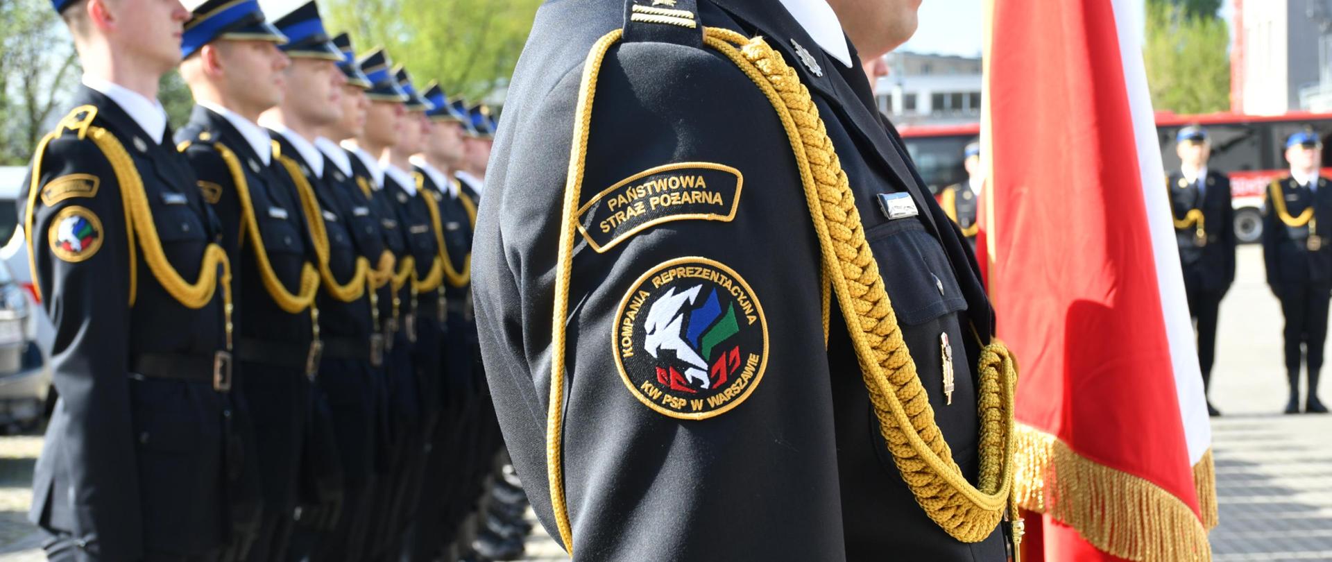 Funkcjonariusze Kompanii Reprezentacyjnej Komendy Wojewódzkiej PSP w Warszawie stoją w dwuszeregu, na pierwszym planie wyostrzenie skierowane na logo Kompanii Reprezentacyjnej znajdujące się na ramieniu jednego ze strażaków
