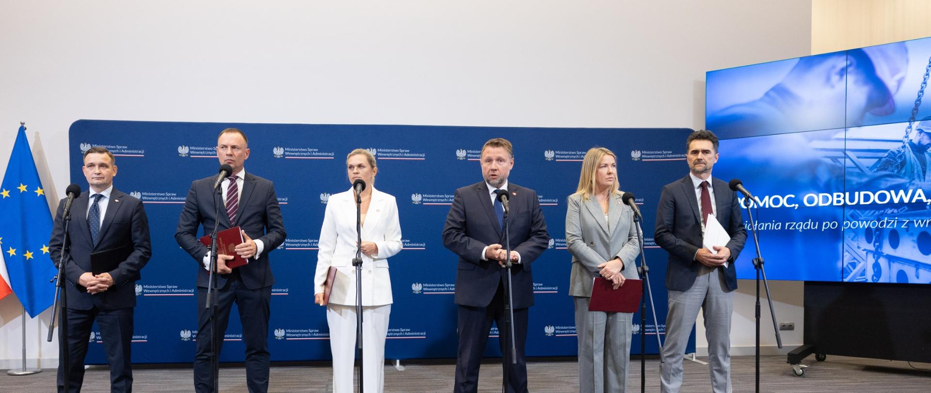 konferencja resortów na temat pomocy po powodzi. Ministrowie stoją za mikrofonami na tle granatowej ścianki z logiem Ministerstwa Spraw Wewnętrznych i Administracji. Po prawej stronie stoi ekran z prezentacją.