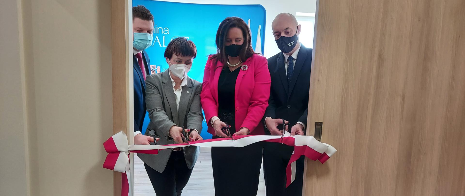 Na zdjęciu cztery osoby, przecinające biało-czerwoną wstęgę z okazji otwarcia klubu Senior Plus w m. Grocholub. Osoby te stoją pomiędzy lewym a prawym skrzydłem drewnianych drzwi. Po lewej stronie stoi wójt gminy Walce, który kieruje nożyczki w stronę wstęgi. Jest to wysoki, postawny mężczyzna, ubrany w ciemny garnitur. Obok niego wicemarszałek województwa, która także przecina wstęgę. Ubrana jest w szary żakiet, ma krótkie brązowe włosy. Kolejną osobą jest poseł na Sejm Porowska, która także przecina wstęgę. Na jej prawym nadgarstku widać białą bransoletkę. Ubrana jest w czarną sukienkę i czerwony żakiet. Patrzy w dół, w kierunku szarfy. Obok stoi wojewoda opolski, który wzrok skupia na poprawnym przecięciu wstęgi. Ubrany jest w ciemny garnitur i granatowy krawat. W tle widać granatową ściankę z napisem Gmina Walce. 