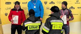 Na zdjęciu widać osoby stojące na scenie oraz podestach podium. Osoby ubrane są w umundurowanie strażackie oraz ubrania sportowe. Za osobami znajdującymi się na scenie znajduje się baner reklamowy koloru żółtego. Osoby znajdujące się na podium trzymają pucharu. Zdjęcie wykonane w dzień. Osoby w umundurowaniu strażackim wręczają nagrody. 