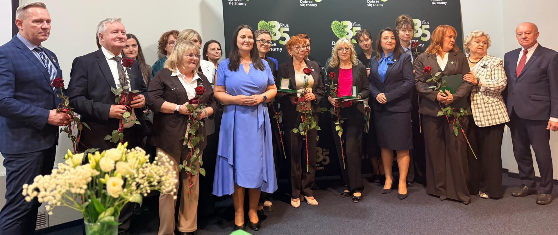 Kilkanaście osób pozuje do zdjęcia na tle ścianki 35-lecia KRUS. Większość z nich trzyma w ręce różę oraz dyplom i etui z wyróżnieniem.